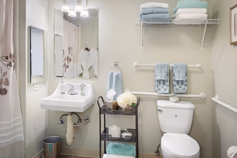 Bright accessible bathroom with a wall-mounted sink, toilet, towel racks and grab bars, shelving stocked with folded towels, and a small cart with decorative items.