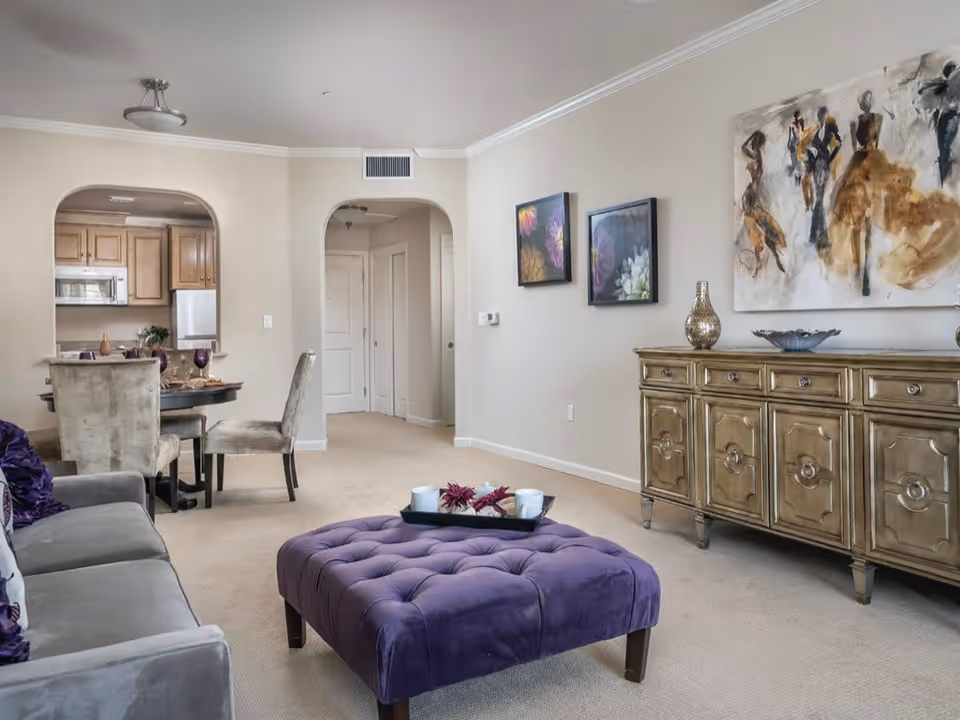 A cozy living room area with a gray sofa adorned with purple pillows, a large purple tufted ottoman with a tray holding two white cups and flowers, a decorative wooden sideboard with vases and a bowl, and three pieces of wall art. In the background, there is a dining area with a round table and upholstered chairs, and a kitchen with wooden cabinets and appliances visible through an arched doorway.