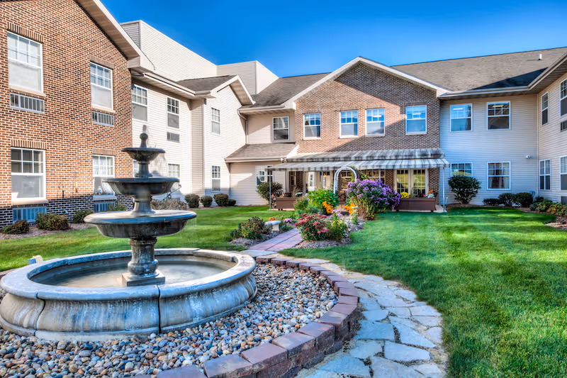 Outdoor courtyard area of a senior living facility with a three-tiered water fountain surrounded by rocks in the foreground, a stone pathway leading to a garden with colorful flowers, and a two-story building with brick and siding in the background under a clear blue sky.