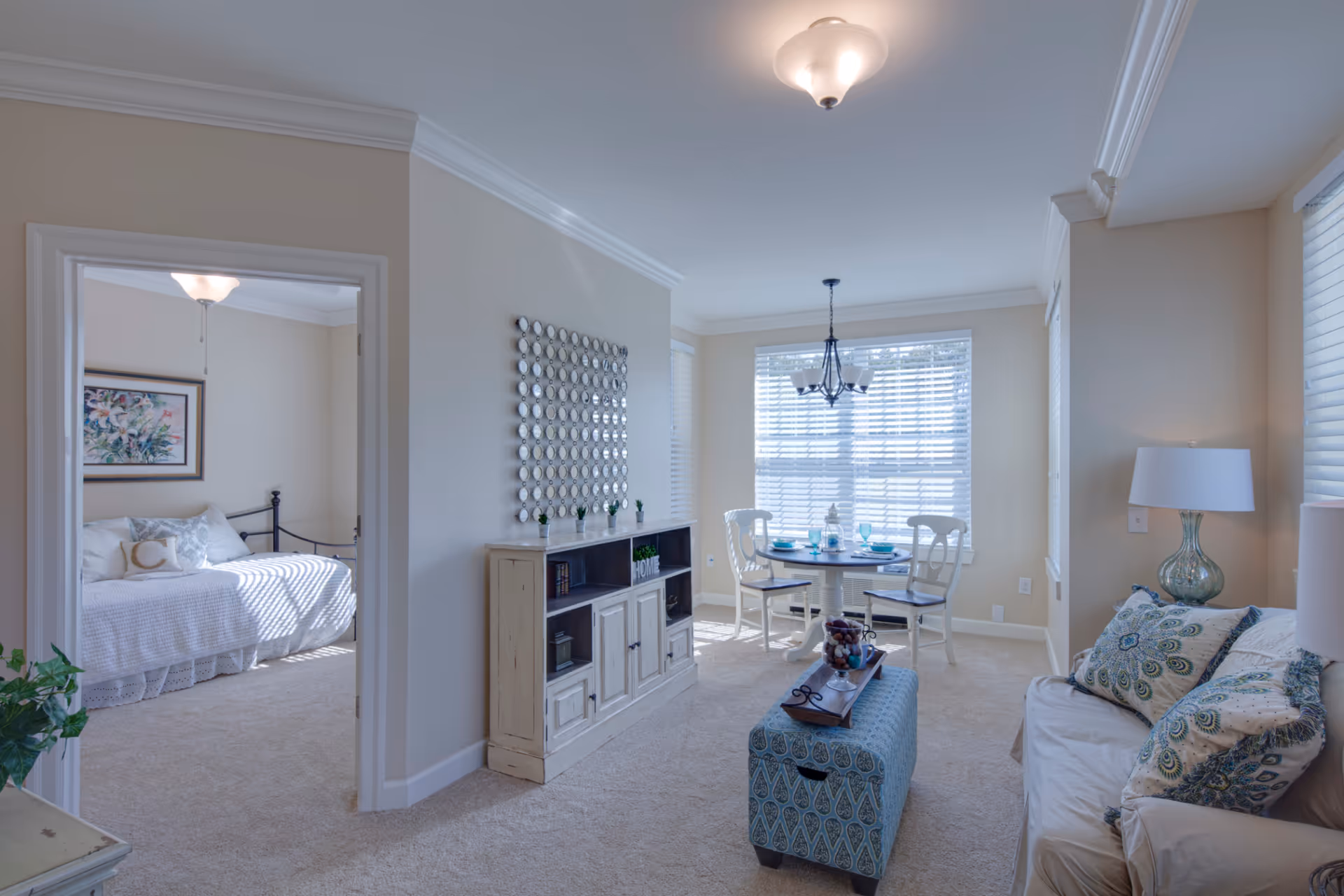 A bright and cozy senior living interior featuring a small dining area with a round table set for two near large windows with blinds. Adjacent to the dining area is a living space with a beige sofa adorned with patterned pillows and a glass table lamp. A decorative cabinet with small plants and a wall mirror is positioned against the wall. To the left, an open doorway reveals a bedroom with a white bedspread and framed artwork above the bed.
