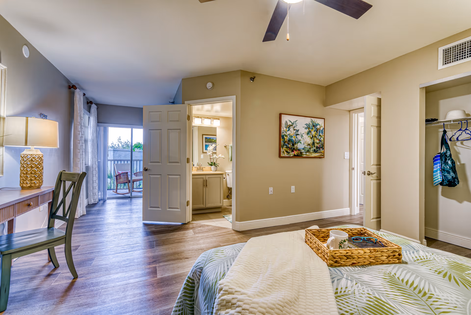 A bright and spacious bedroom in a senior living facility with a bed covered in a green and white leaf-patterned bedspread and a beige blanket. A wicker tray with a mug and sunglasses sits on the bed. To the right is an open closet with hanging clothes and bags. A door leads to a bathroom with a vanity, sink, and toilet visible. A sliding glass door at the far end opens to a balcony with chairs. A wooden chair and desk with a lamp are on the left side of the room. The room has wood flooring and neutral-colored walls with a ceiling fan overhead.