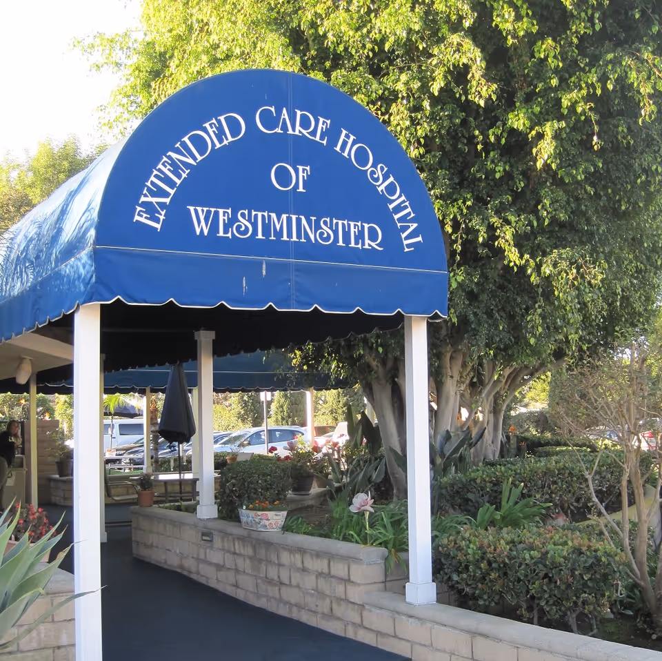 Outdoor covered walkway with a blue awning that reads 'Extended Care Hospital of Westminster', surrounded by greenery and plants with parked cars visible in the background.
