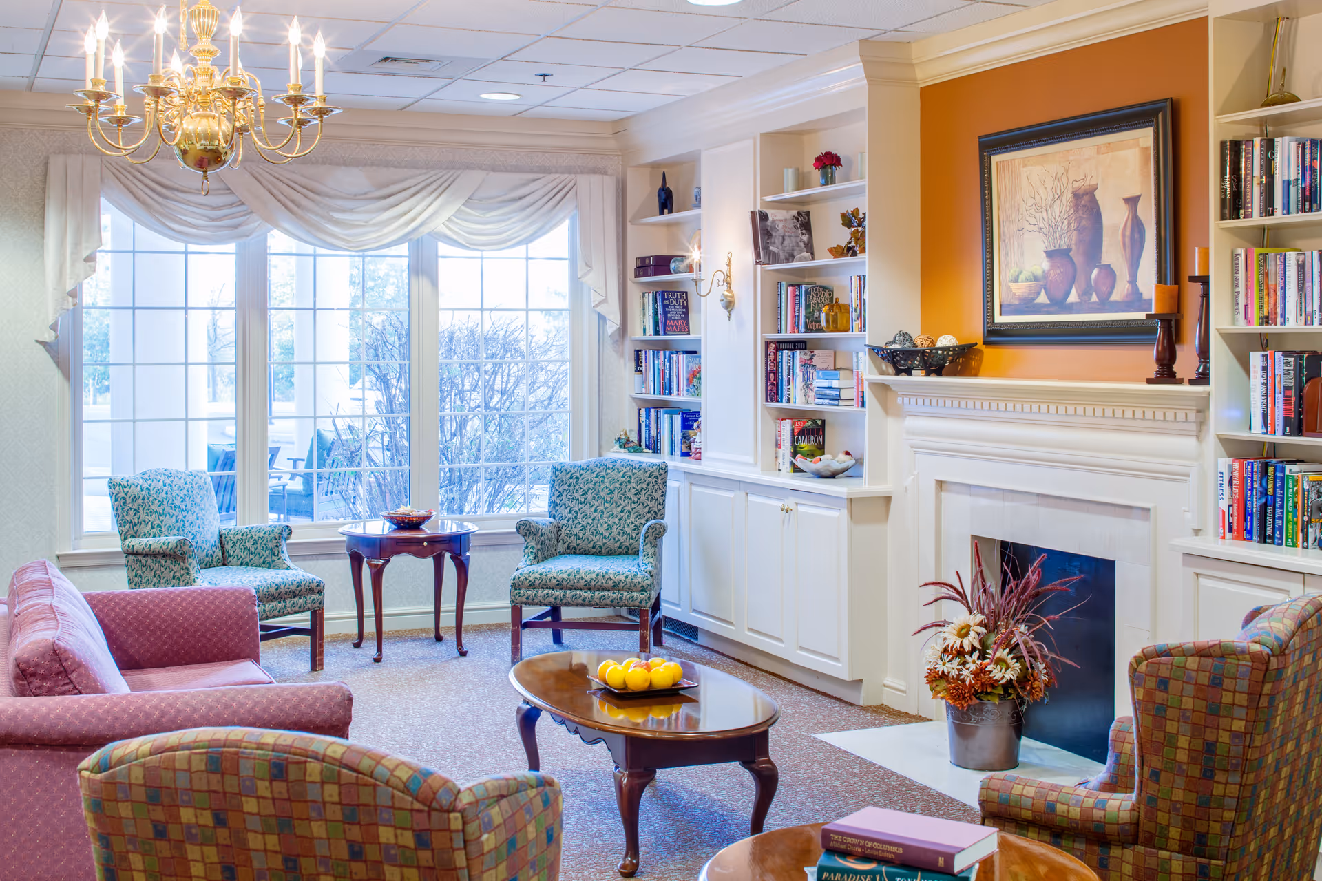 A cozy living room with patterned armchairs and a pink sofa arranged around wooden coffee tables. There is a fireplace with a floral arrangement in front, built-in white bookshelves filled with books and decorative items, and a large window with draped curtains letting in natural light. A chandelier hangs from the ceiling.
