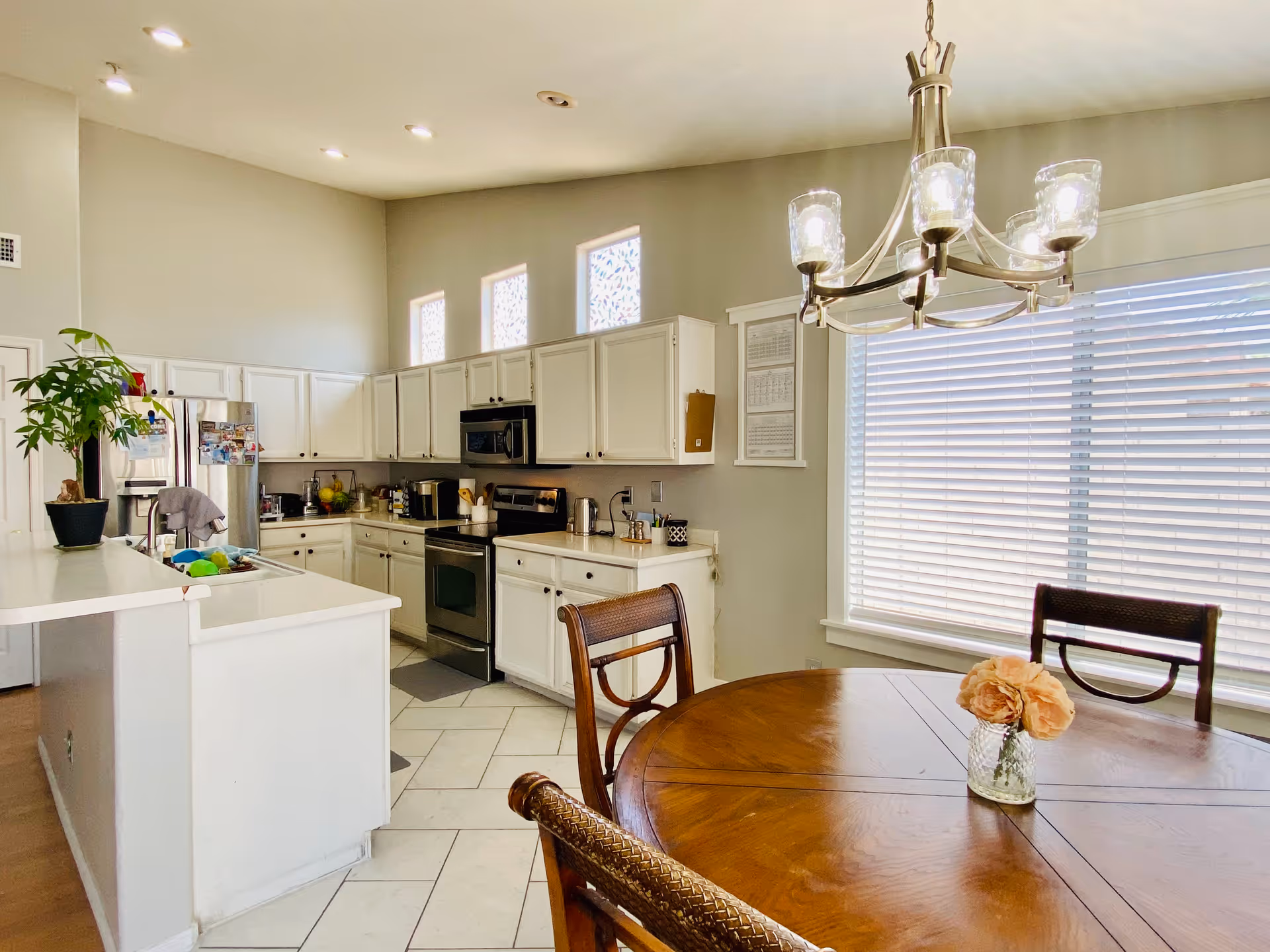 Bright kitchen and dining area with white cabinets, stainless steel appliances, a round wooden dining table with chairs, a chandelier overhead, and a large window with blinds letting in natural light.