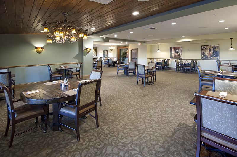 Spacious dining room with multiple wooden tables and chairs arranged neatly on a patterned carpet. The room features a wooden ceiling with a decorative chandelier, soft wall sconces, and framed artwork on the walls. The lighting is warm and inviting, creating a comfortable atmosphere.