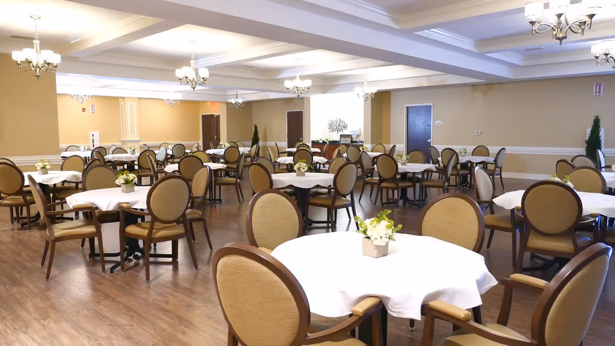 A spacious dining room with multiple round tables covered with white tablecloths, each surrounded by beige cushioned chairs with wooden frames. Small floral centerpieces decorate each table. The room has wooden flooring, beige walls, and several chandeliers hanging from a coffered ceiling providing warm lighting.