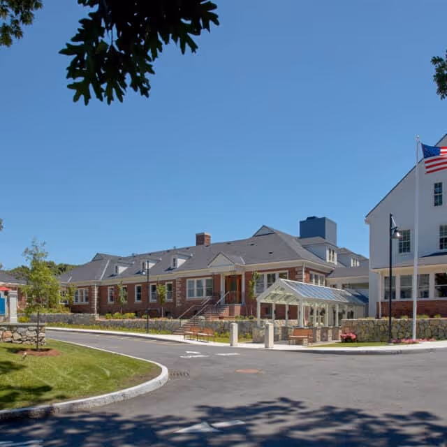 Exterior view of Forestdale Park Senior Living facility showing a large brick building with multiple windows, a glass-enclosed entrance area, an American flag on a flagpole, and a curved driveway surrounded by landscaped greenery under a clear blue sky.
