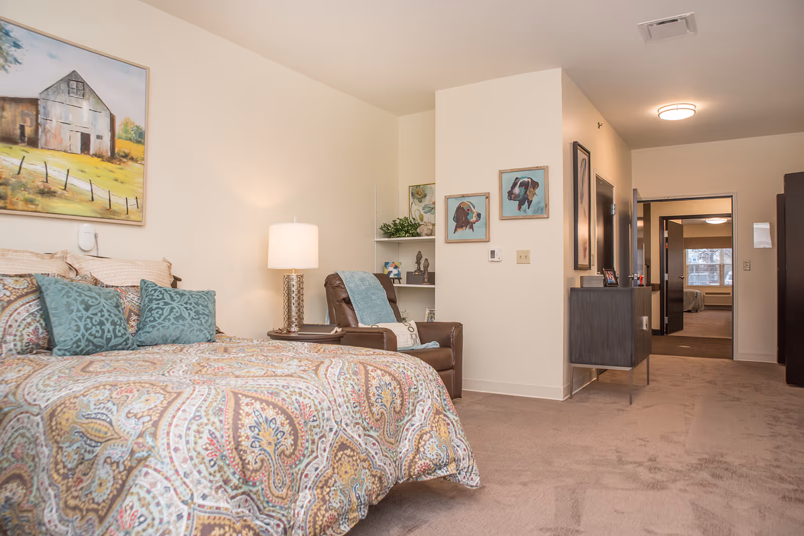 Bright furnished bedroom with a patterned bedspread, armchair and lamp, and an open doorway leading to another room.