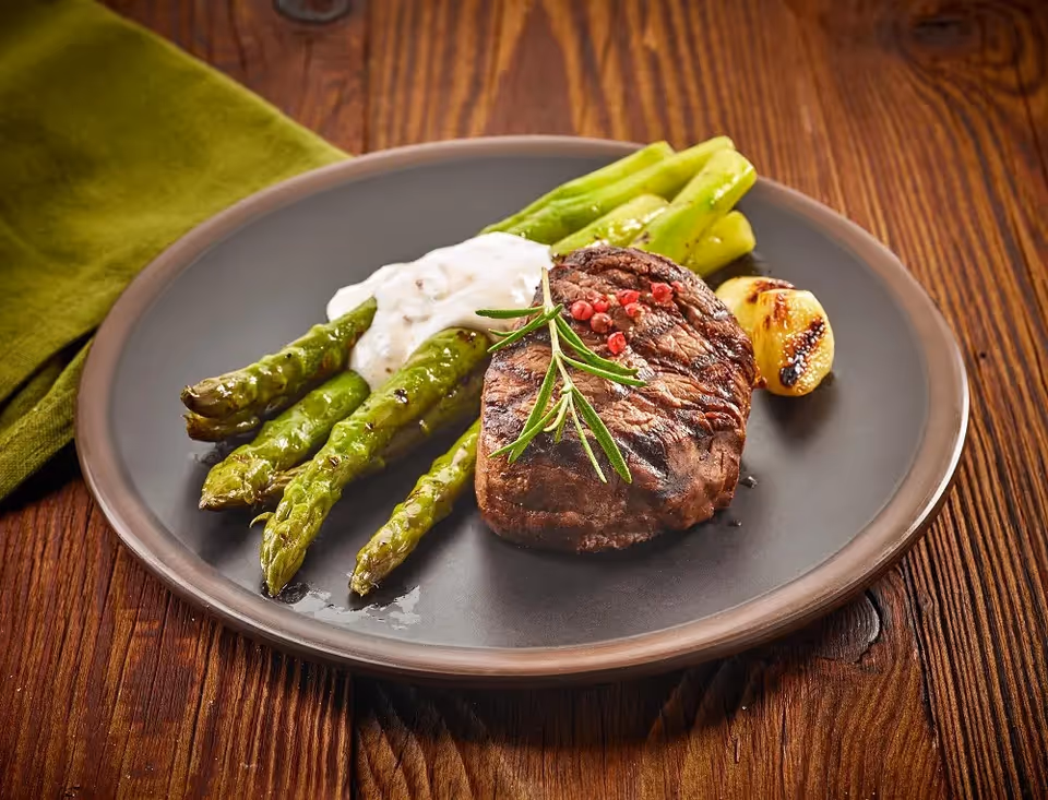 A plate with grilled asparagus, a grilled steak garnished with a sprig of rosemary and pink peppercorns, a dollop of white sauce, and a grilled lemon wedge on a wooden table with a green napkin.