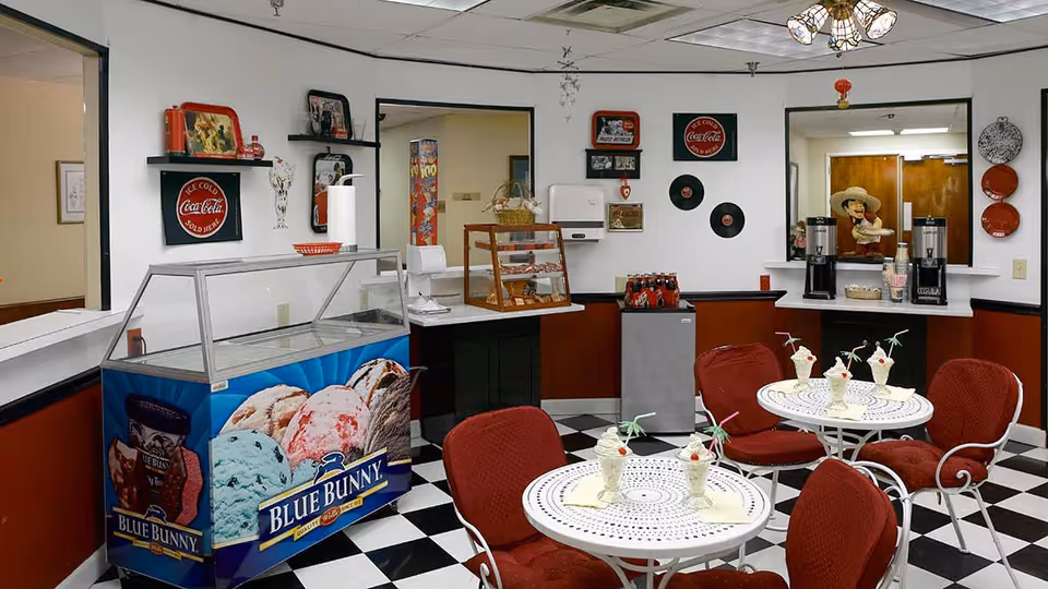 A retro-style ice cream parlor area with black and white checkered floor, red cushioned chairs, and white round tables. Each table has several ice cream sundaes with cherries and small umbrellas. There is a Blue Bunny ice cream freezer, a small display case with baked goods, and coffee dispensers on a counter. The walls are decorated with vintage Coca-Cola signs, vinyl records, and red plates.