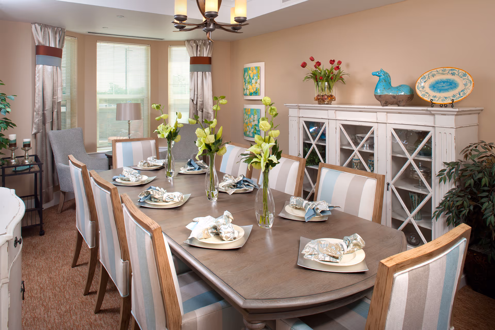 A well-decorated dining room with a long wooden table set for eight people. Each place setting has a plate with a folded napkin. Three vases with green flowers are placed along the center of the table. The room features large windows with blinds and curtains, a white cabinet with glass doors displaying dishes, and decorative items including a blue horse figurine, a plate, and a vase with red tulips. There are also two framed floral paintings on the wall and a potted plant in the corner.