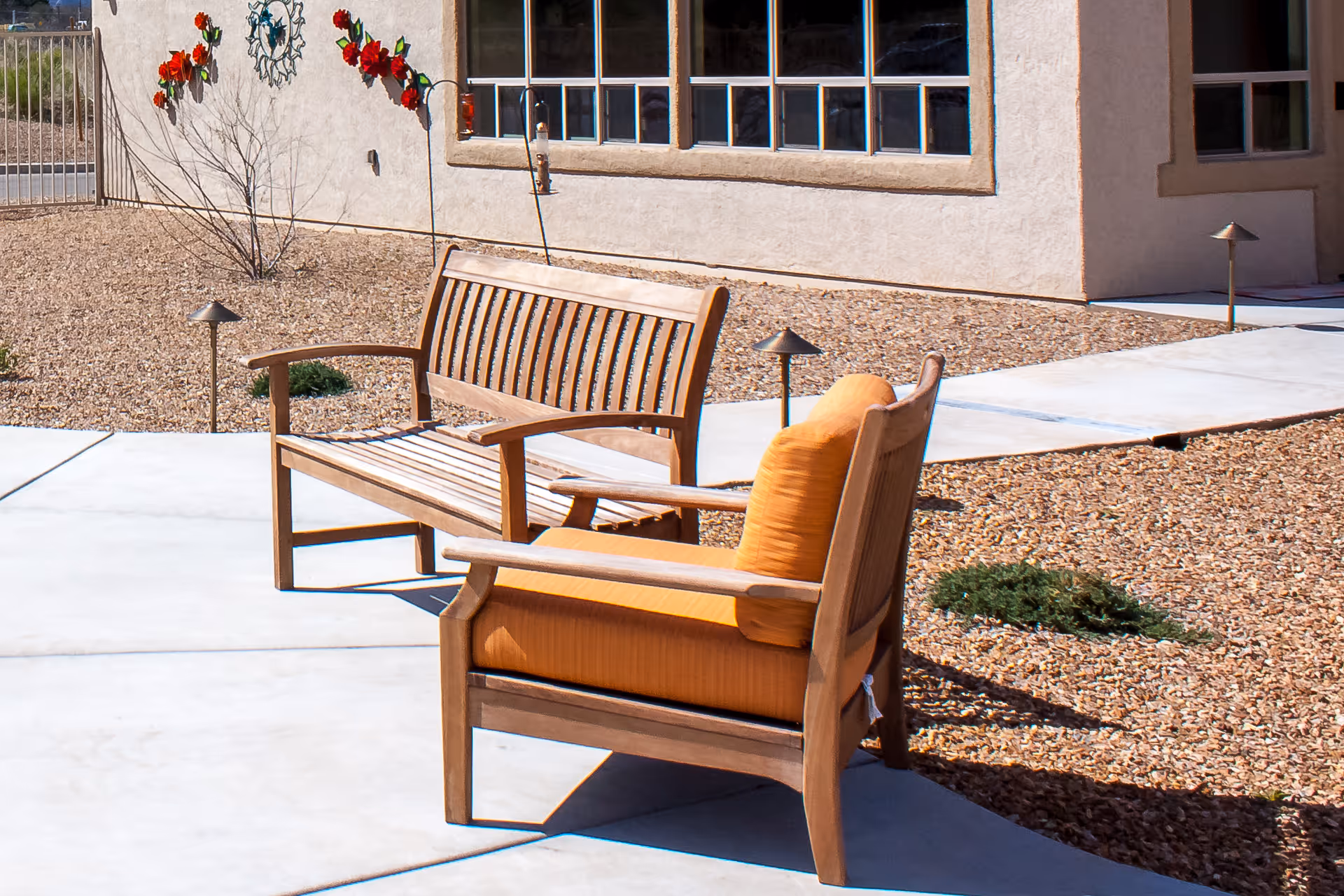 Outdoor seating area with a wooden bench and a wooden chair with orange cushions on a concrete patio next to a building with windows. The ground around the patio is covered with small rocks and some sparse plants. Decorative red flower wall art is mounted on the building's exterior wall.