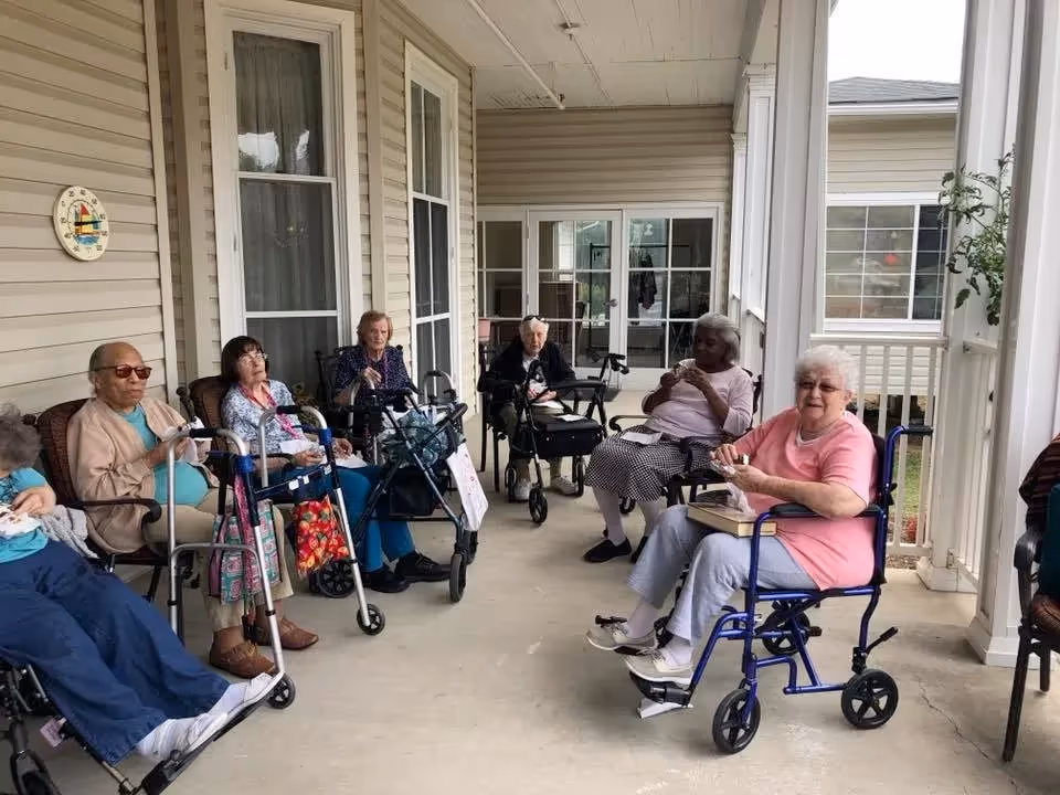 Several elderly residents seated with walkers and wheelchairs on a covered porch at an assisted living facility.
