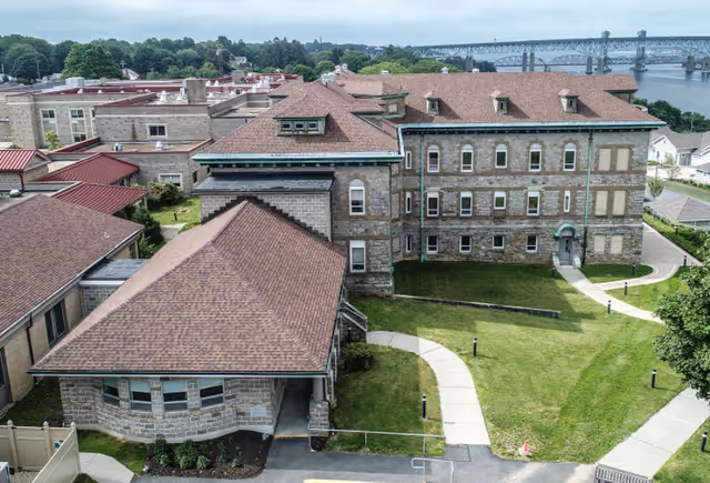 Aerial view of a senior living facility named Fairview showing multiple connected stone buildings with brown shingled roofs, green lawns, paved walkways, and a large bridge over a body of water in the background.
