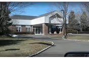 Front entrance of a single-story brick and white nursing facility with a covered drop-off, circular driveway, and leafless trees.