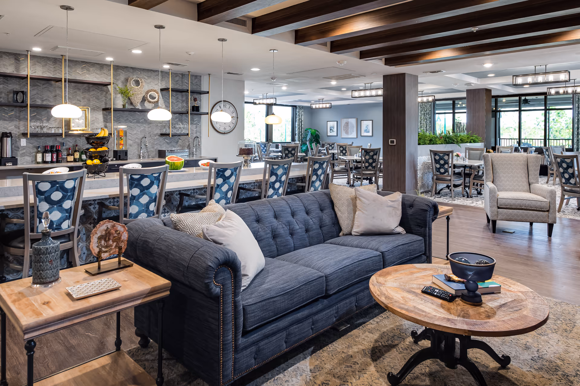 A spacious and modern common area in a senior living facility featuring a dark blue tufted sofa with pillows, a wooden coffee table with decorative items and a remote control, and a side table with a lamp and decorative stone. Behind the sofa is a long counter with blue patterned chairs, pendant lights, and a bar area with bottles and fruit. In the background, there are multiple dining tables and chairs, large windows letting in natural light, and ceiling lights with wooden beams overhead.