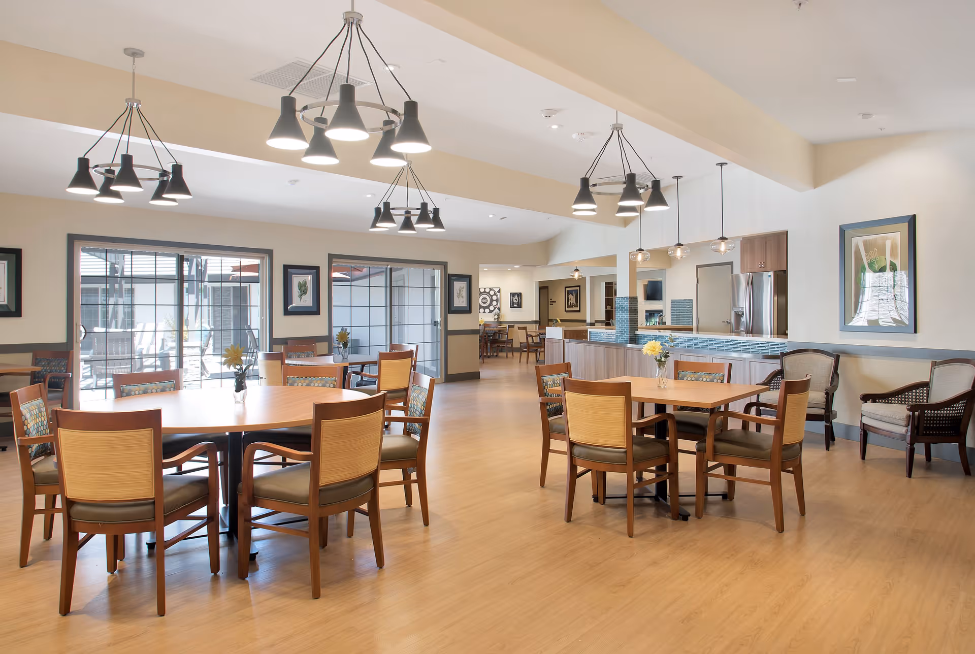 A bright and spacious dining area in an assisted living facility with wooden tables and chairs arranged neatly. The room features modern pendant lighting, large windows with views of an outdoor patio, and a kitchen area with stainless steel appliances and a blue tile backsplash. The walls are decorated with framed artwork and the floor is light wood.