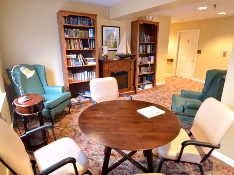 A cozy common area with a round wooden table surrounded by four cream-colored chairs with black armrests. There are two green armchairs near a small wooden side table with a lamp. In the background, there are two wooden bookshelves filled with books and a decorative fireplace with a model sailboat on top. The room has patterned carpet and light yellow walls with a hallway and a white door visible.