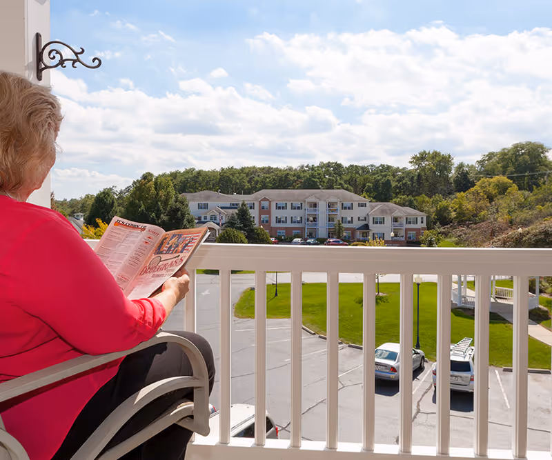 An elderly woman in a pink top sitting on a balcony chair reading a newspaper, overlooking a parking lot, green lawn, and a multi-story residential building with trees in the background under a partly cloudy sky.