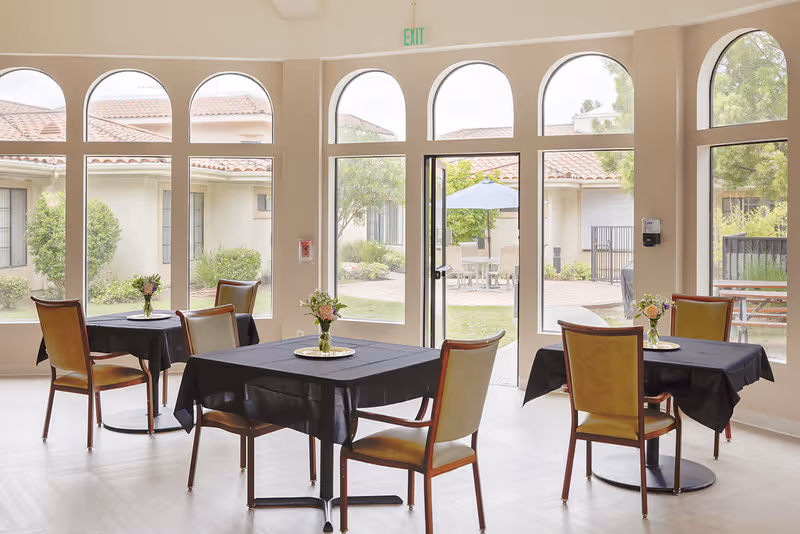 A bright dining area with several tables covered in black tablecloths, each adorned with a small vase of flowers. The room features large arched windows and a glass door that opens to an outdoor patio with seating and an umbrella. The walls are light-colored, and the floor has a light wood finish.