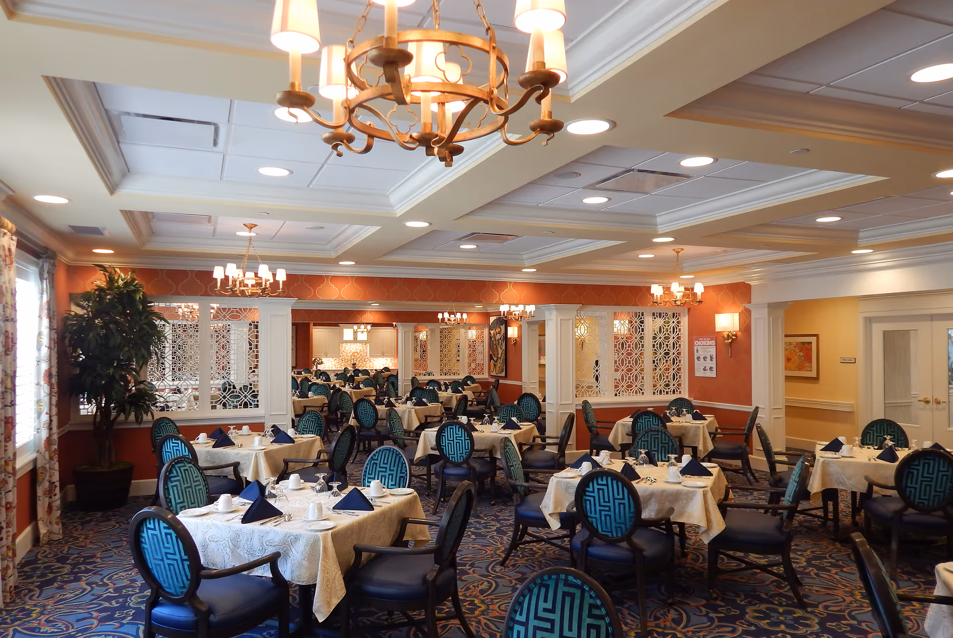 A spacious dining room in an assisted living facility with multiple round tables covered with white tablecloths, each set with white cups, plates, silverware, and navy blue folded napkins. The chairs have dark wood frames and blue patterned upholstery. The room features ornate chandeliers, recessed lighting, decorative white partitions, and patterned carpet. There is a large plant near the window with floral curtains and warm-colored walls.