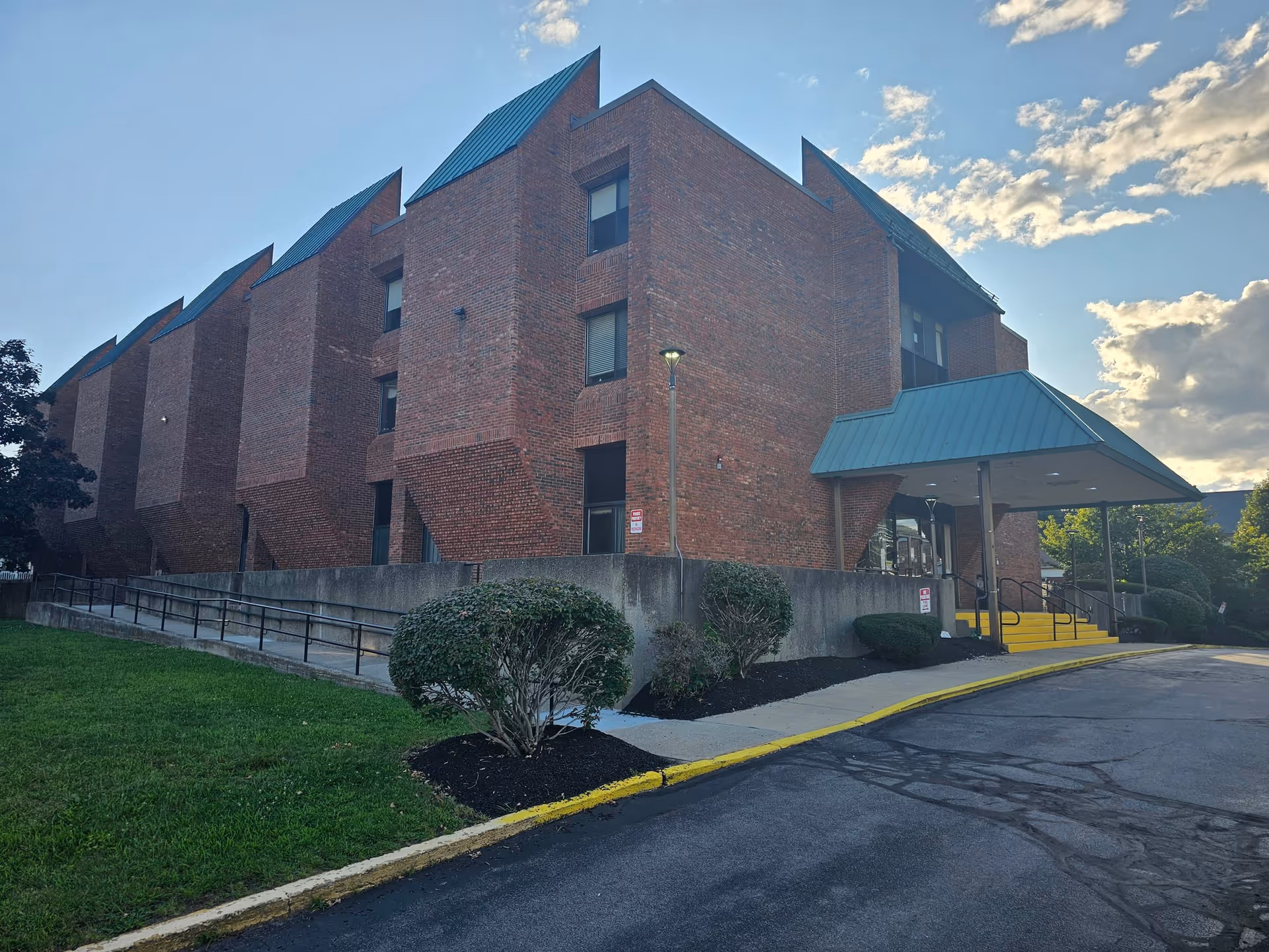 Front exterior of a multi-story brick senior living building with a green metal entrance awning, wheelchair ramp, and landscaped shrubs under a blue sky.