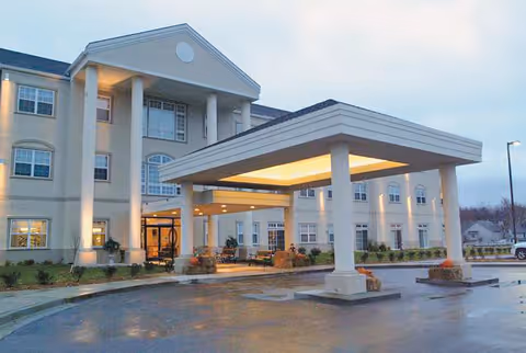 Exterior view of a large, three-story retirement community building with a covered entrance supported by columns. The building is illuminated with warm lights during dusk, and there are some small plants and pumpkins near the entrance.