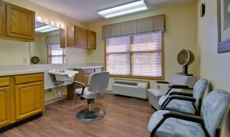 Interior salon/styling room with a barber chair and wash sink, wooden cabinets, waiting chairs, and a window with blinds.