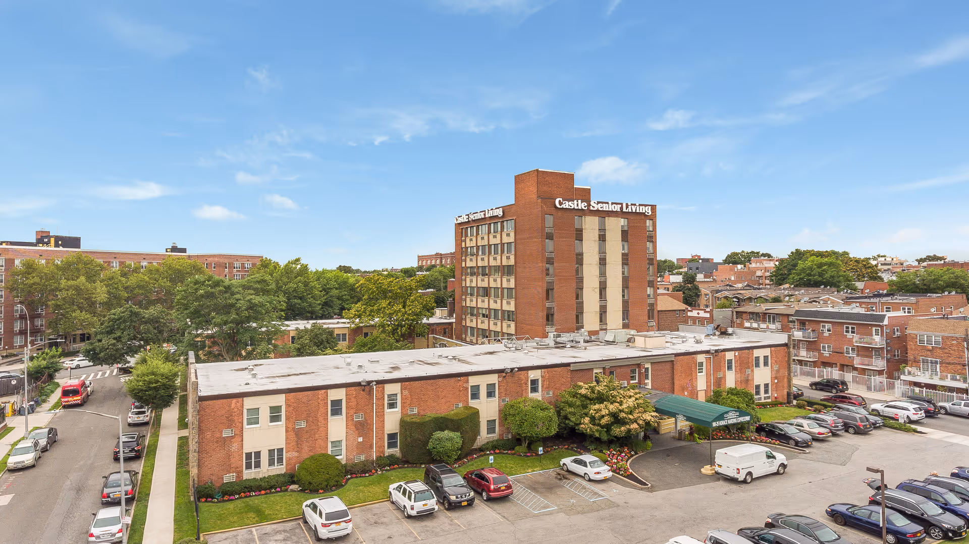 Exterior view of a multi-story brick senior living building labeled "Castle Senior Living" with an adjacent parking lot and surrounding neighborhood under a blue sky.