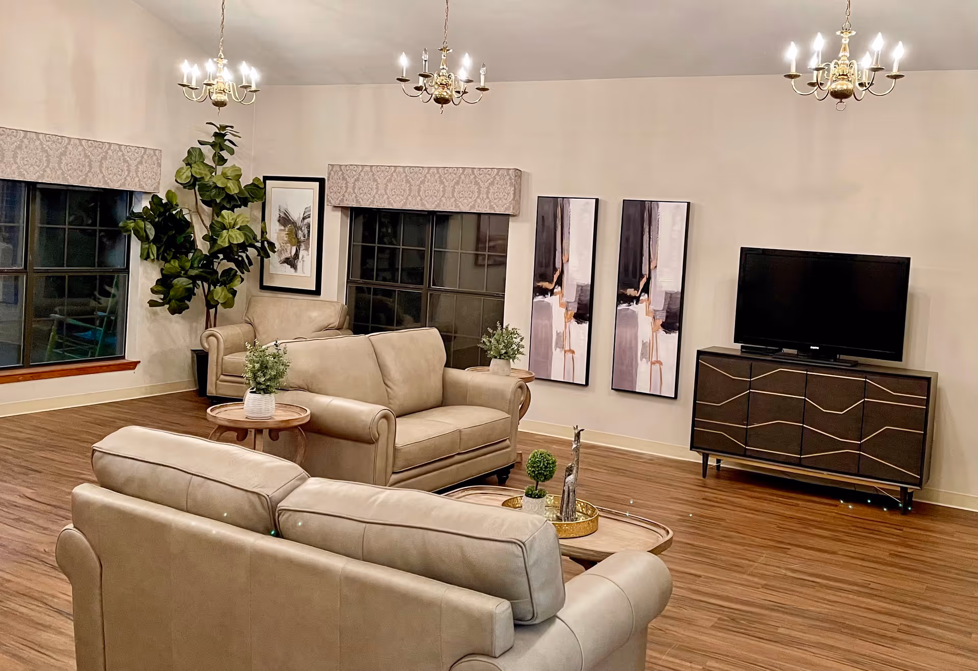 A cozy living room with beige leather sofas arranged around a wooden coffee table with decorative plants. The room features wooden flooring, large windows with patterned valances, abstract wall art, a flat-screen TV on a modern dark wooden cabinet, and three brass chandeliers hanging from the ceiling.