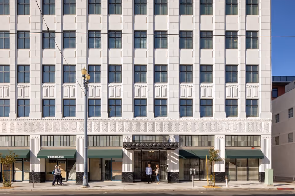 Front exterior view of a multi-story white building with many windows and green awnings over the ground floor entrances. Two people are walking on the sidewalk in front of the building, and two others are standing near the entrance. There is a streetlamp and parking meters visible on the sidewalk.