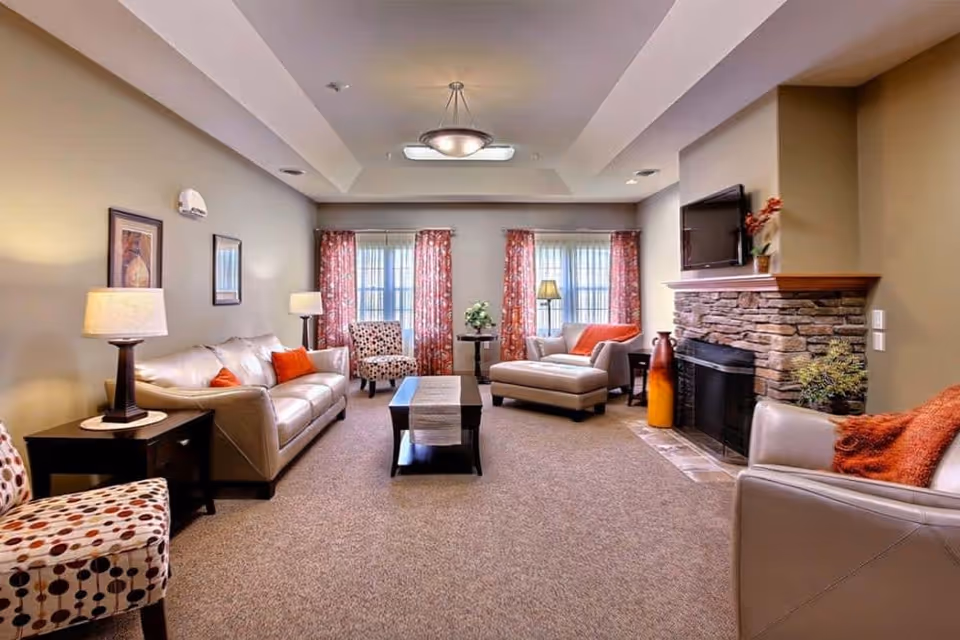 A bright living room with sofas, armchairs, a coffee table, windows with patterned curtains and a stone fireplace with a wall-mounted TV.