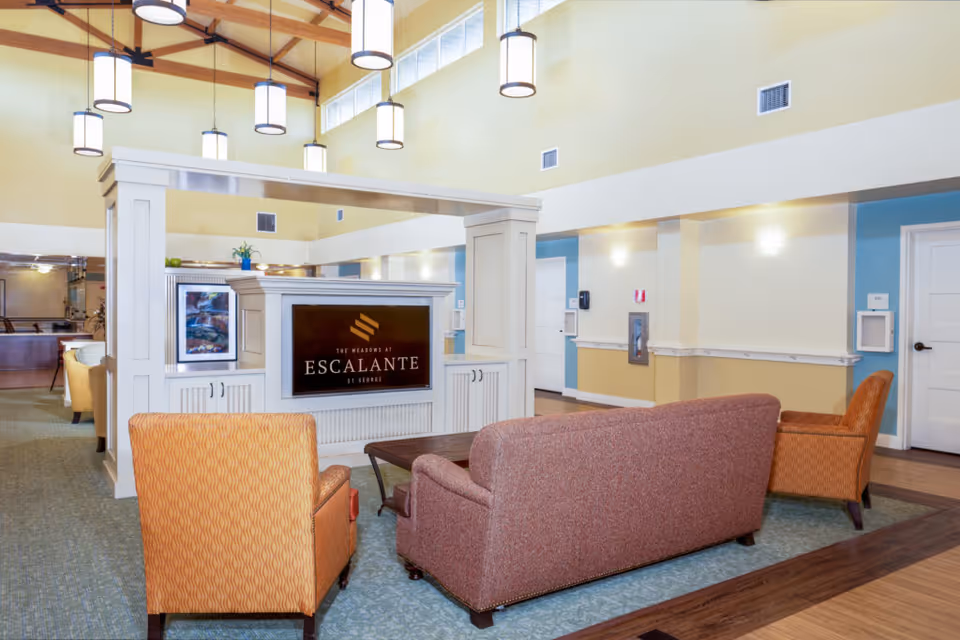 Bright senior living lounge with sofas and armchairs facing a central partition displaying 'Escalante' signage under hanging pendant lights.