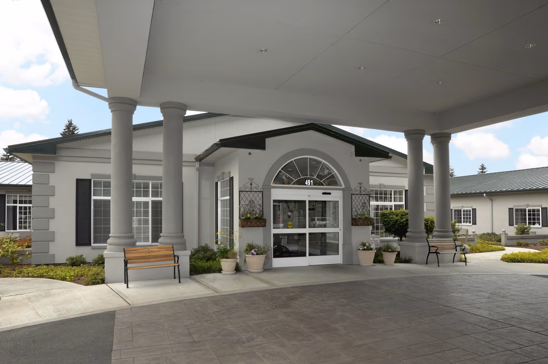Covered main entrance of a senior living facility with columns, benches, potted plants, and sliding glass doors labeled 491.