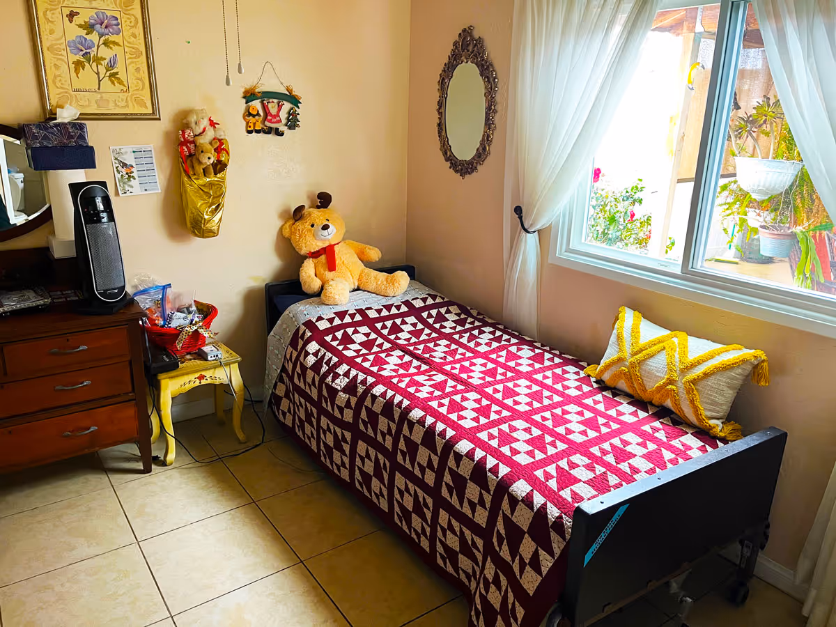 Small bedroom with a single bed covered by a red-and-white quilt, a large teddy bear, dresser, side table, and a window with sheer curtains.