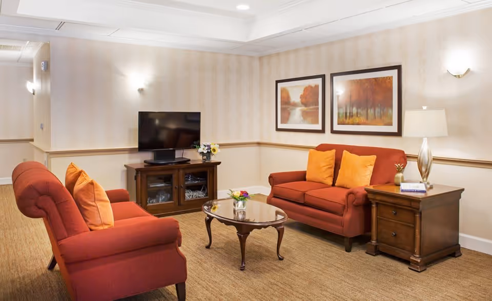 A cozy living room with two red sofas adorned with orange and yellow pillows, a wooden coffee table with a small flower arrangement, a wooden TV stand with a flat-screen TV, and two framed autumn landscape paintings on the wall. A wooden side table with a lamp and a small decorative item is next to one of the sofas. The room has beige wallpaper and carpeted flooring.