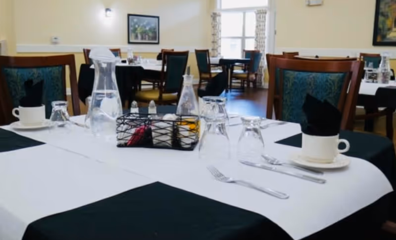 A dining room table set with white and black tablecloths, glass water pitchers, upside-down drinking glasses, white cups with black napkins, silverware, and a condiment basket. In the background, there are more tables and chairs, a window with curtains, and framed artwork on the walls.