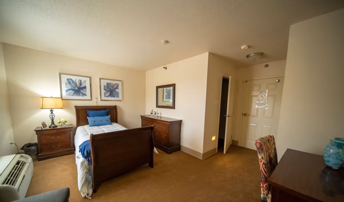 A cozy bedroom with a single wooden bed featuring white bedding and blue pillows. There is a wooden nightstand with a lamp and small decorative items on the left side of the bed. Above the bed, two framed blue floral artworks are hung on the wall. To the right of the bed, there is a wooden dresser with a mirror above it. The room has beige walls and carpet, a closed white door, and a patterned chair next to a wooden desk on the right side.