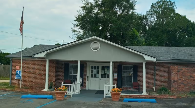 Front exterior view of a single-story brick building with a covered entrance supported by white columns. There are two large flower pots with orange flowers on either side of the entrance ramp. An American flag is flying on a flagpole to the left, and there are two handicap parking spaces in front of the building. Trees and greenery are visible in the background.
