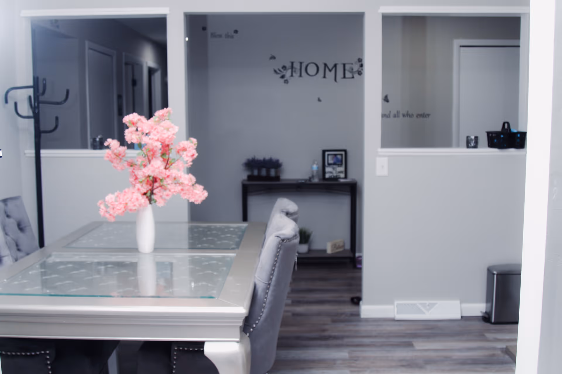Interior view of a residential assisted living facility dining area with a glass-top table and gray upholstered chairs. A white vase with pink flowers is placed on the table. The background shows a hallway and a small table against the wall with decorative items and a framed picture. The wall has a decorative text that reads 'HOME'.