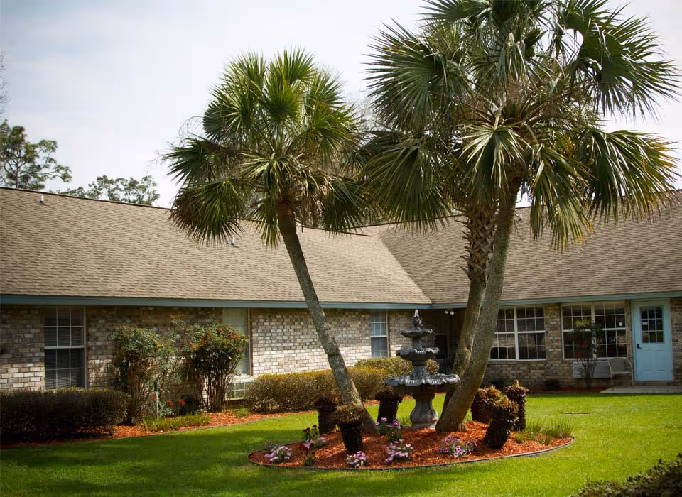 Front exterior of a brick senior living building with palm trees, a tiered fountain, and a manicured lawn.