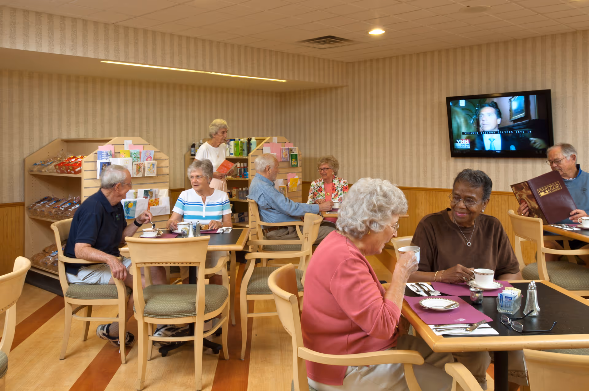 A group of elderly people sitting and conversing at tables in a dining area. Some are drinking from cups, while one person is reading a menu. Shelves with snacks and greeting cards are visible in the background, along with a wall-mounted television showing a news program.