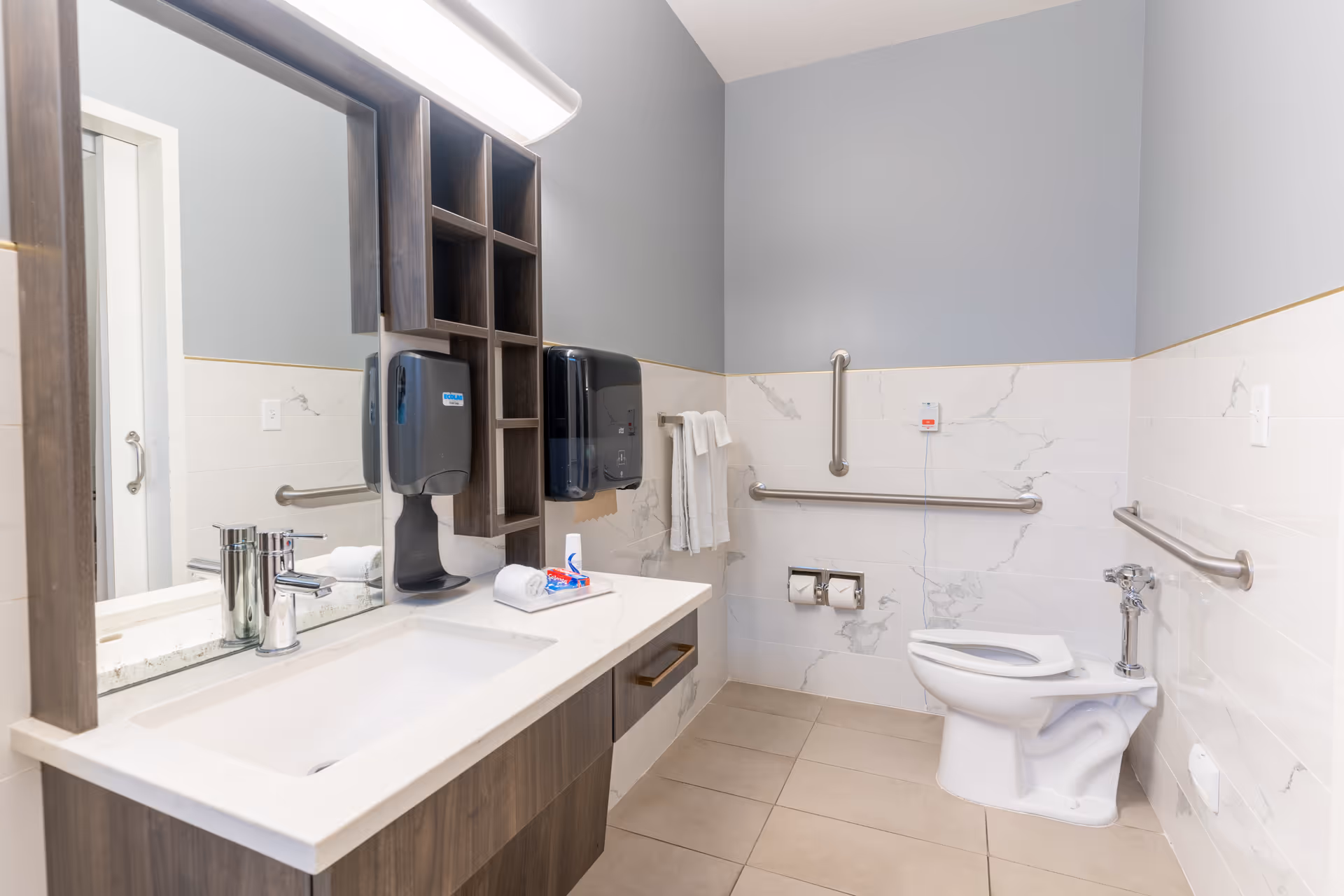 A clean, modern bathroom with a white sink and countertop, a large mirror above the sink, and wooden cabinetry. The bathroom features accessibility grab bars around the toilet area, a wall-mounted soap dispenser, paper towel dispenser, and a towel rack with white towels. The walls are tiled halfway up with a marble pattern and painted gray above.
