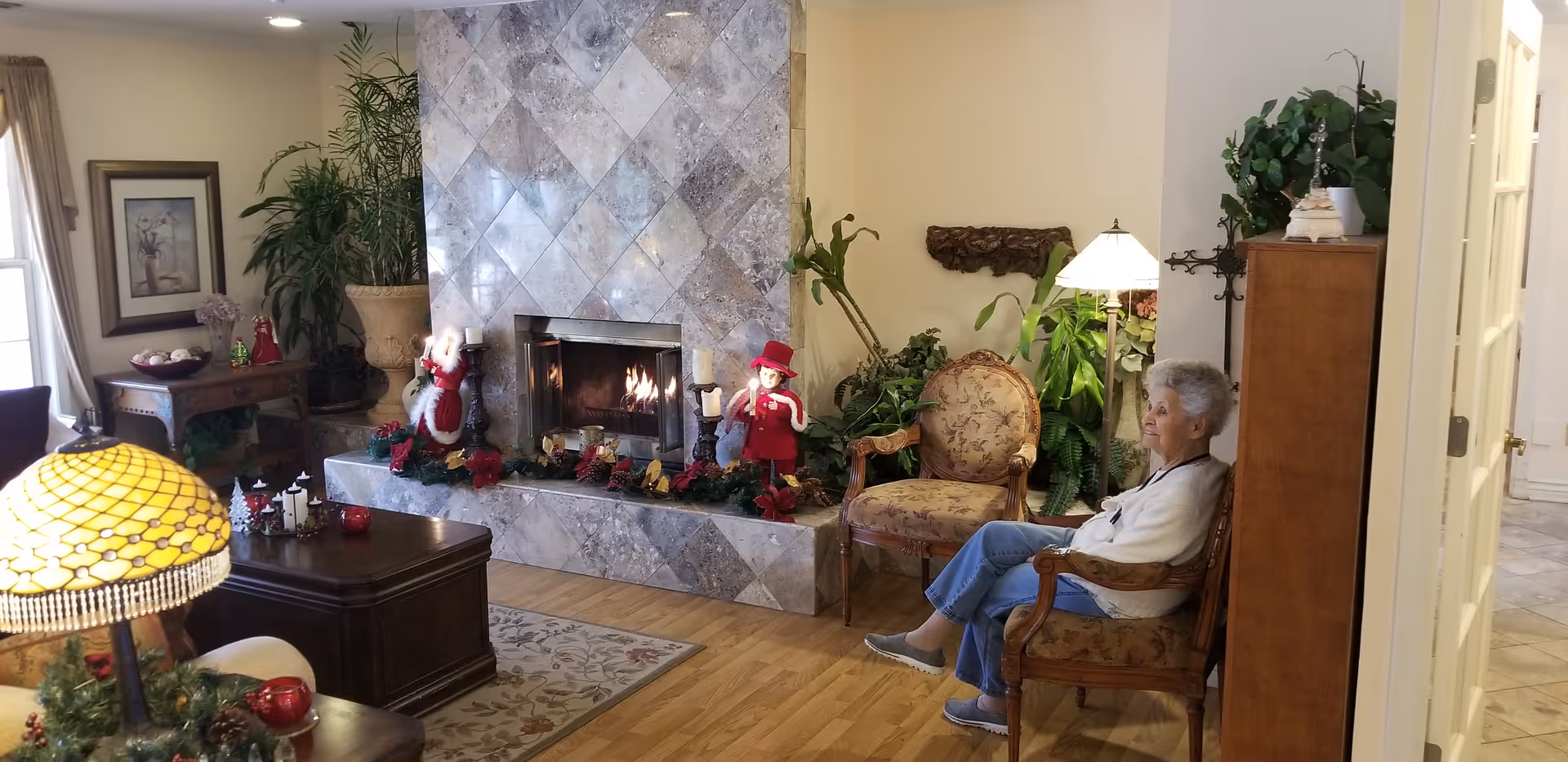A cozy living room with a tiled fireplace decorated with Christmas ornaments and garlands. An elderly woman is seated on a floral upholstered wooden chair near the fireplace. The room features wooden flooring, a patterned rug, a wooden coffee table, a Tiffany-style lamp, various plants, and framed artwork on the walls.