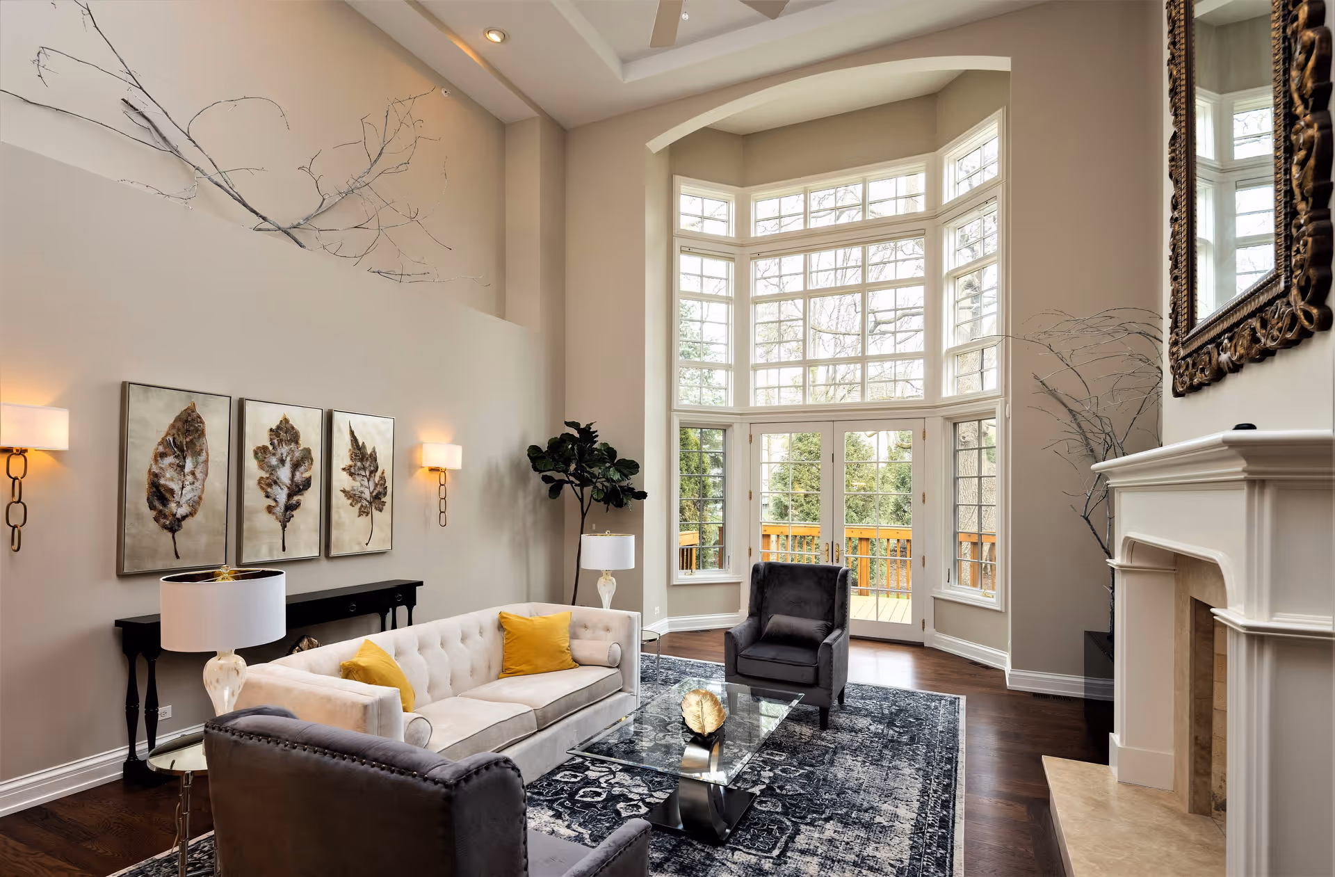 A spacious living room with high ceilings and large bay windows letting in natural light. The room features a beige tufted sofa with yellow pillows, two gray armchairs, a glass coffee table with a decorative gold leaf, a dark patterned rug, a white fireplace with a large ornate mirror above it, and wall art depicting three leaves. There are also two wall sconces, a floor lamp, a side table, and a potted plant near the windows.