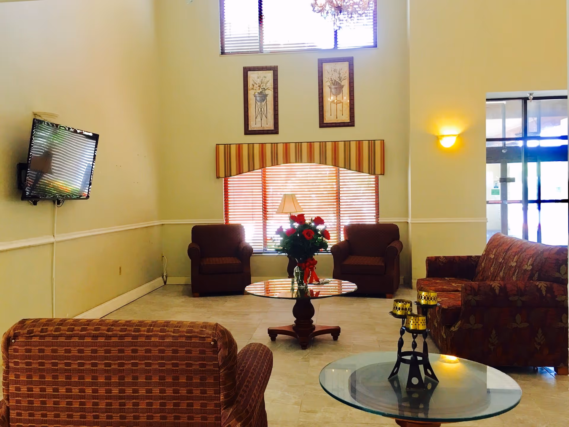 A cozy living room area with a high ceiling and large windows covered with blinds. The room features two armchairs and a sofa with patterned upholstery, a glass coffee table with a floral centerpiece, and a wall-mounted flat-screen TV. There are two framed floral artworks on the wall above the window, and a glass door entrance is visible on the right side.