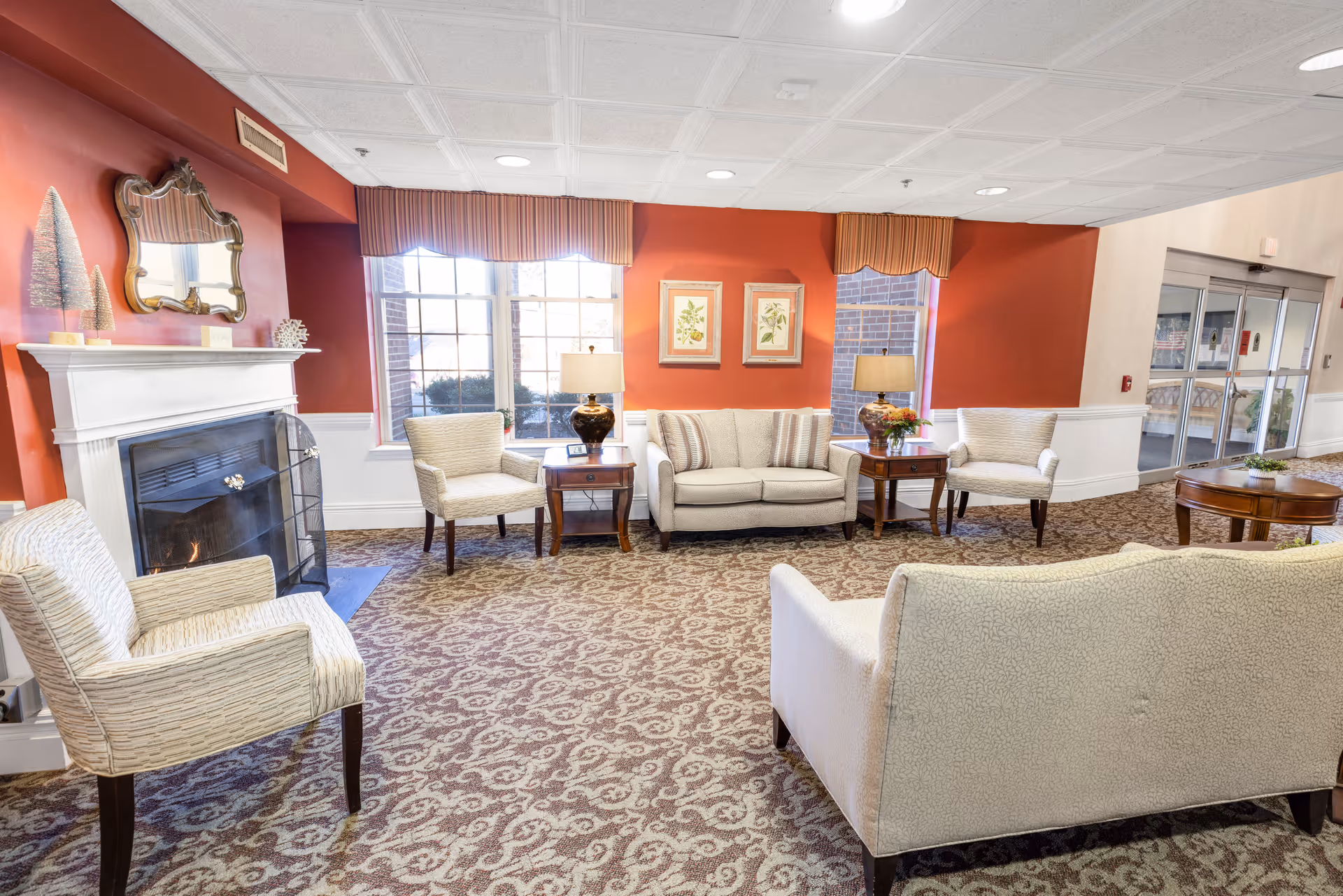 A cozy living room area with patterned carpet, a white fireplace with a fire burning, and several upholstered chairs and sofas arranged around wooden side tables with lamps. The walls are painted red with white wainscoting, and there are large windows with striped valances letting in natural light. Two framed botanical prints hang on the wall above a sofa. A glass door entrance is visible in the background.