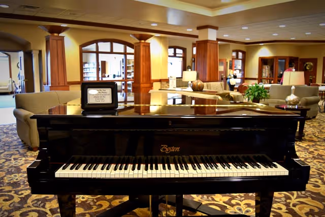 A black Boston grand piano in the foreground of a spacious, well-lit common area with patterned carpet, beige sofas, wooden columns, and large windows in the background.