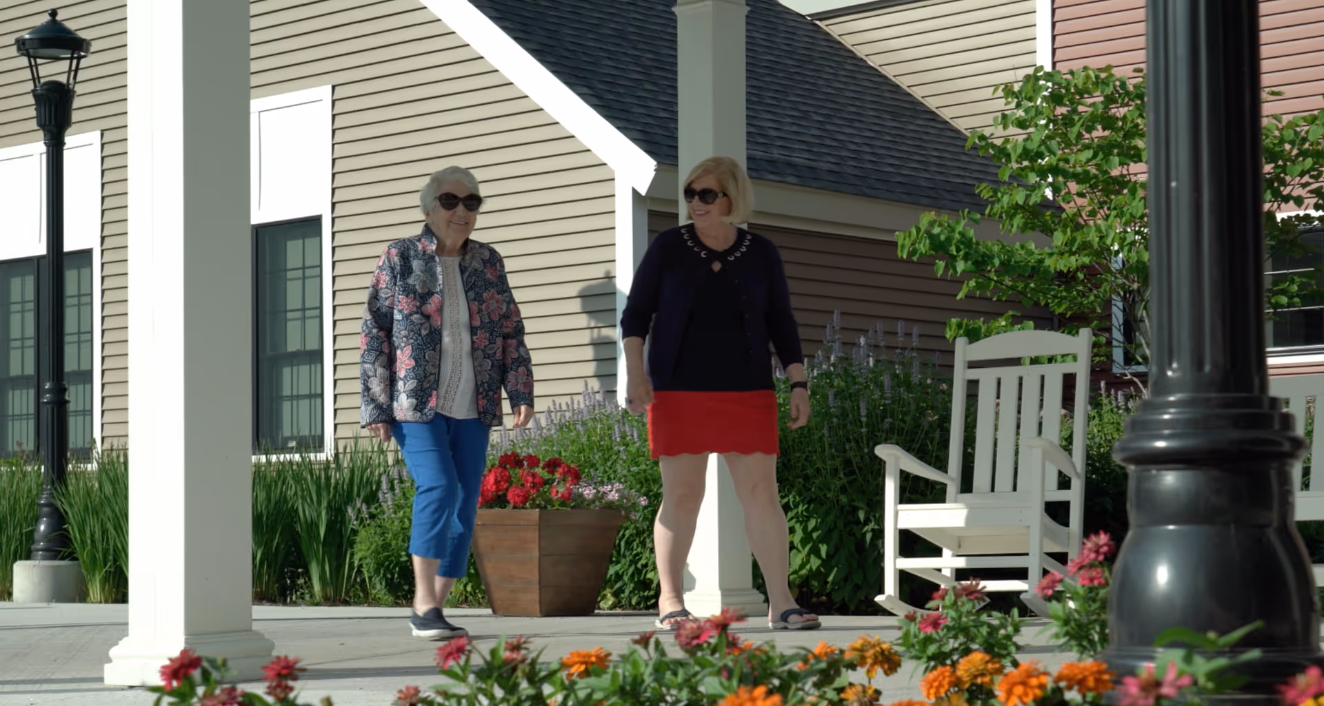 Two women walking outside near a building with beige siding, surrounded by greenery and colorful flowers. There is a white rocking chair and a black lamp post nearby.