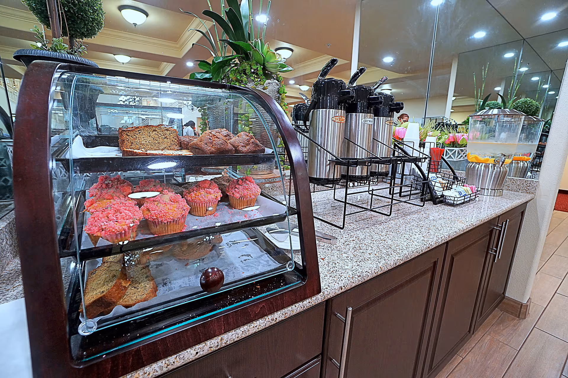 A countertop display case with muffins and slices of bread, next to several stainless steel coffee dispensers and a beverage dispenser with fruit-infused water, set in a well-lit interior space with plants and mirrors in the background.